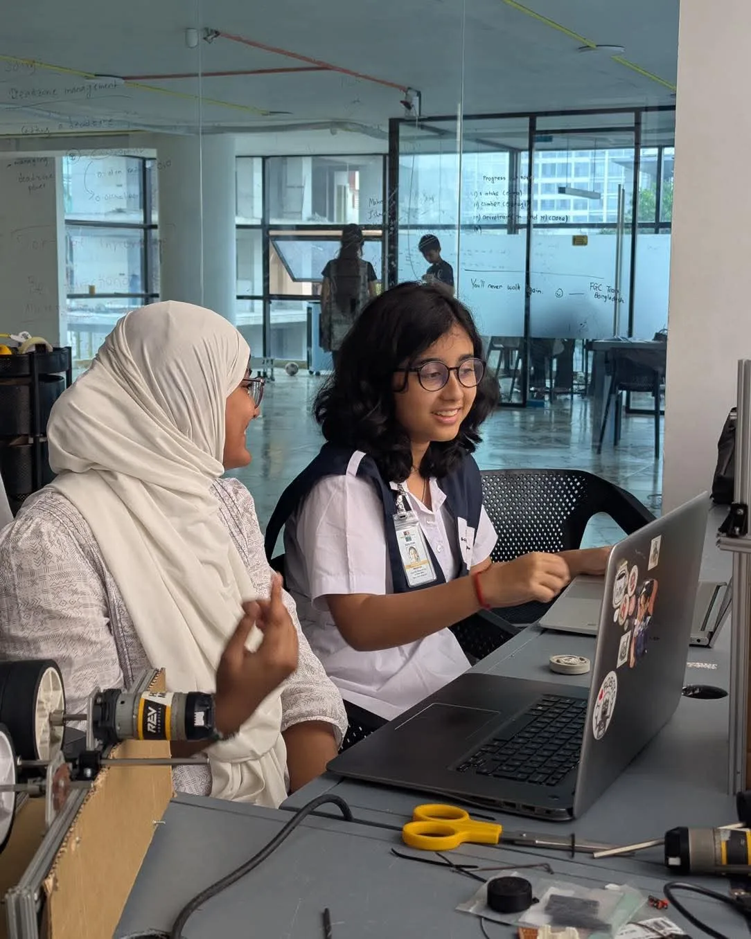 A mentor in a headscarf working through code on a laptop beside a smiling student at a workbench in The Tech Academy lab.