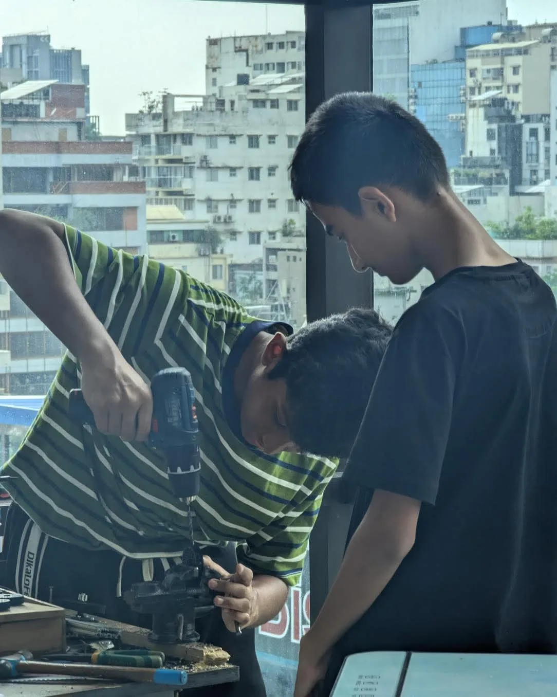 Two students using a power drill to build a metal robotics frame at a window overlooking Dhaka.