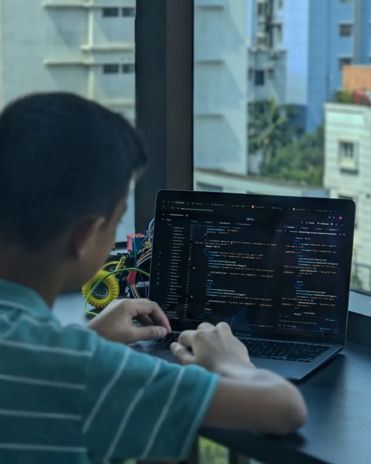 A student coding on a laptop next to a wired robotics car, looking out over Dhaka rooftops.